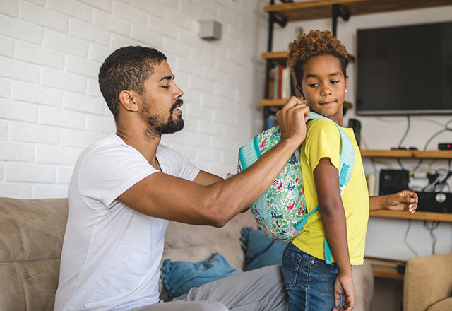 a father helps his daughter put on her backpack at home before school