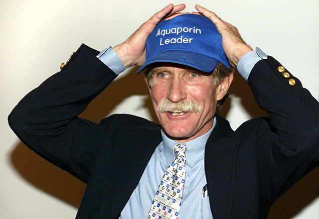 Peter Agre holds his head with both hands in an expression of delighted surprise; he is wearing a hat that reads 