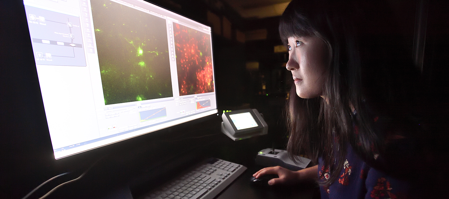 A grad student works on a computer in a dark room, her face illuminated by the screen.