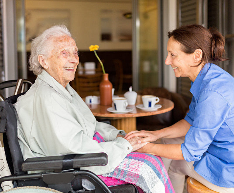 An elderly woman and her adult daughter share laughter together while seated at a table.